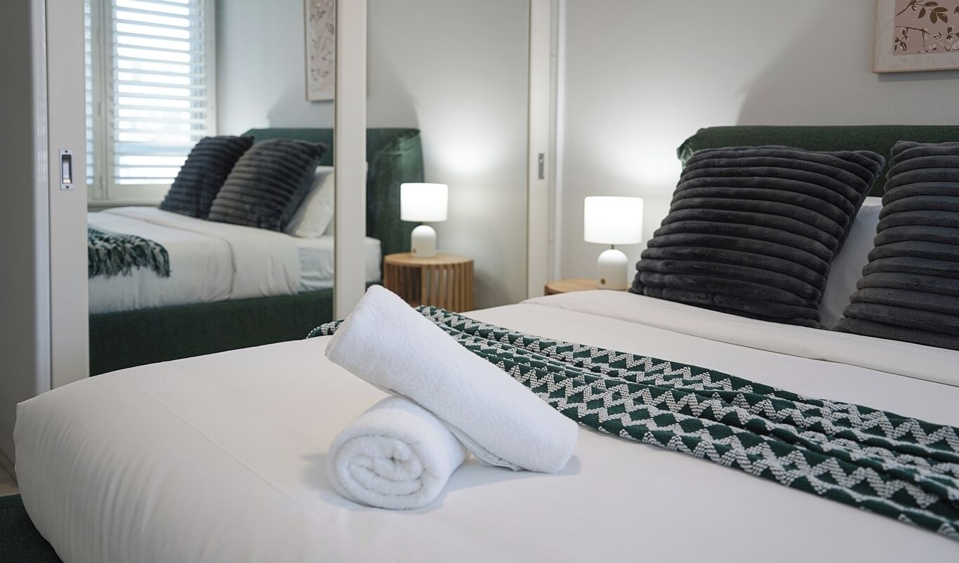 Cama bem arrumada com manta branca e verde estampada, toalha branca enrolada e duas luminárias de cabeceira.