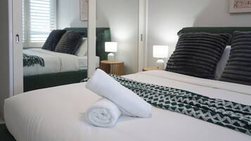 Cama bem arrumada com manta branca e verde estampada, toalha branca enrolada e duas luminárias de cabeceira.