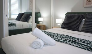 A neatly made bed with a white and green patterned blanket, a rolled white towel, and two bedside lamps. - KozyGuru | Mosman | Serene Boutique Apt Retreat (Mosman)