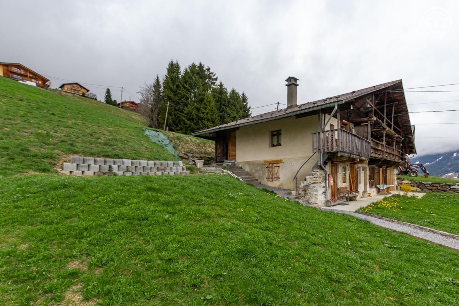 A hillside house with a stone staircase, a wooden balcony, and a mountain backdrop.
