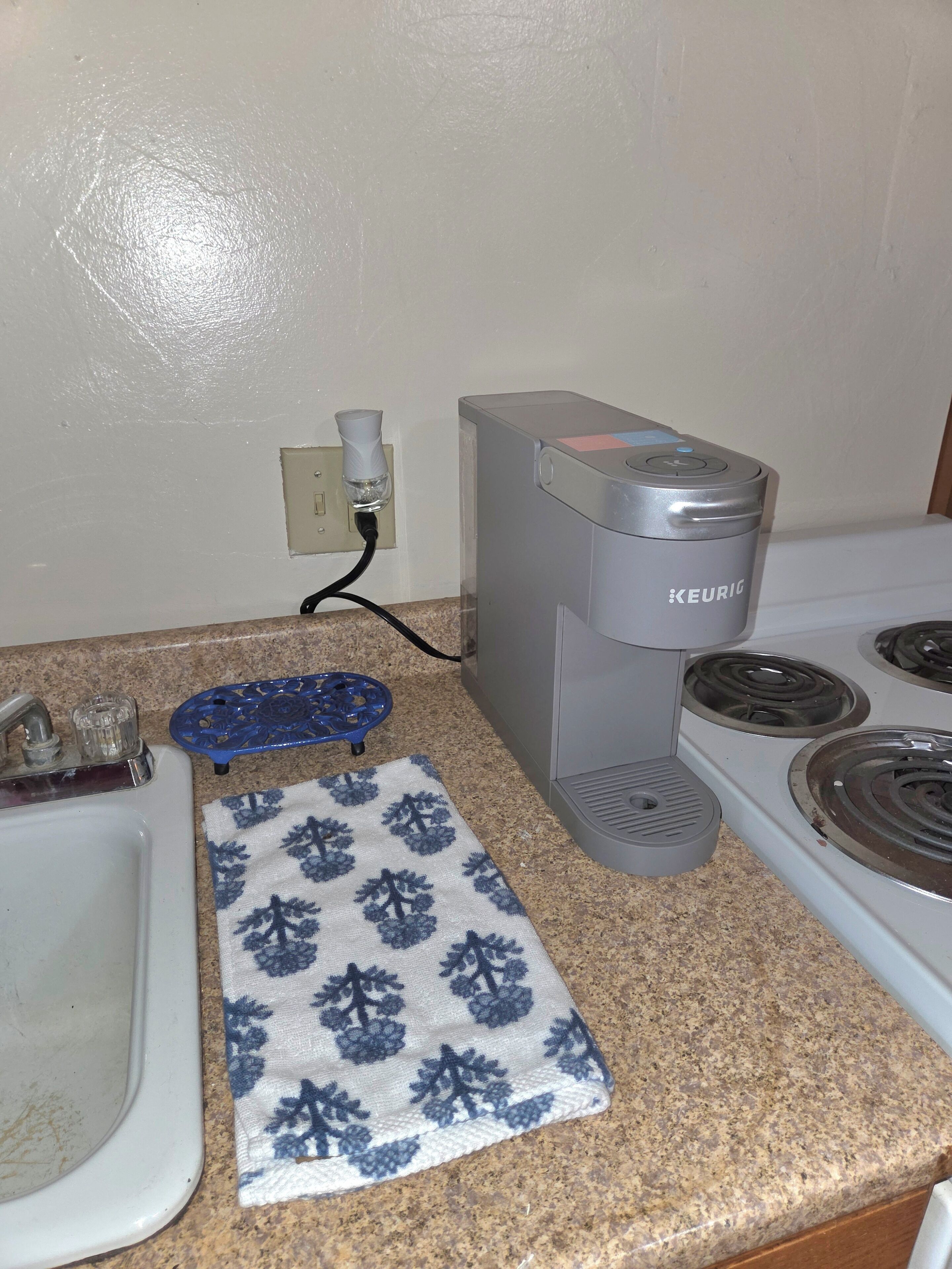 Een keuken met een Keurig koffiezetapparaat, een spoelbak, een fornuis en een geruit doekje op het aanrecht.