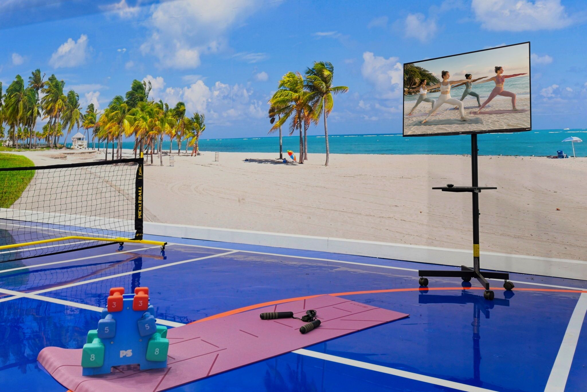 Een strandvolleybalveld met een net, trainingsapparatuur en een scherm waarop een yogales wordt vertoond.