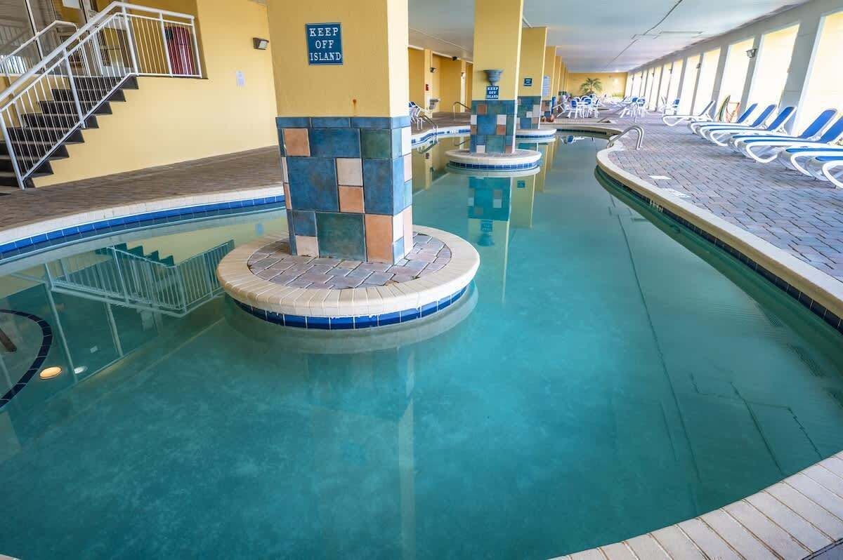 An indoor swimming pool with a 'KEEP OFF ISLAND' sign, a staircase, and lounge chairs along the poolside.