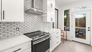 Cucina con mobili bianchi, zoccolo in mattoni, cappa in acciaio inossidabile e una porta scorrevole in vetro che dà su un'area esterna.