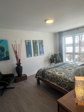 A modern bedroom with a large bed, a desk with a lamp, a chair, a vase with plants, and abstract wall art.
