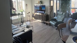 Ein modernes Wohnzimmer mit Flachbildfernseher, großem Fenster und Essbereich mit Tisch und Stühlen.