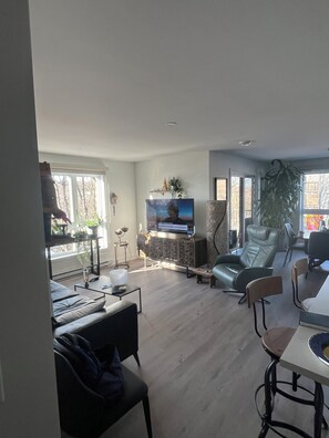 A modern living room with a flat-screen TV, a large window, and a dining area with a table and chairs.