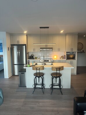 A modern kitchen with stainless steel appliances, wooden bar stools, and light-colored cabinetry.