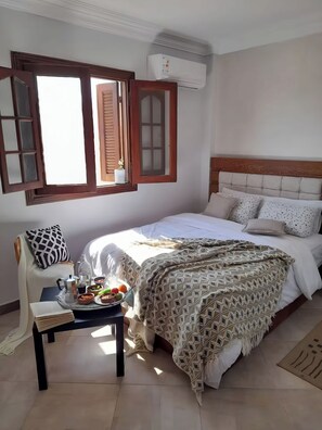 A bedroom with a bed, a bedside table with a tray of food, a chair, and a window with shutters. - Prime Location with Stunning View: Opposite the Museum of Egyptian Civilization, (Fustat)