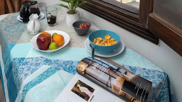Ein Zimmer mit Frühstückstisch, einem Fenster mit Blick auf Gebäude, einer Topfpflanze und einer Zeitschrift.