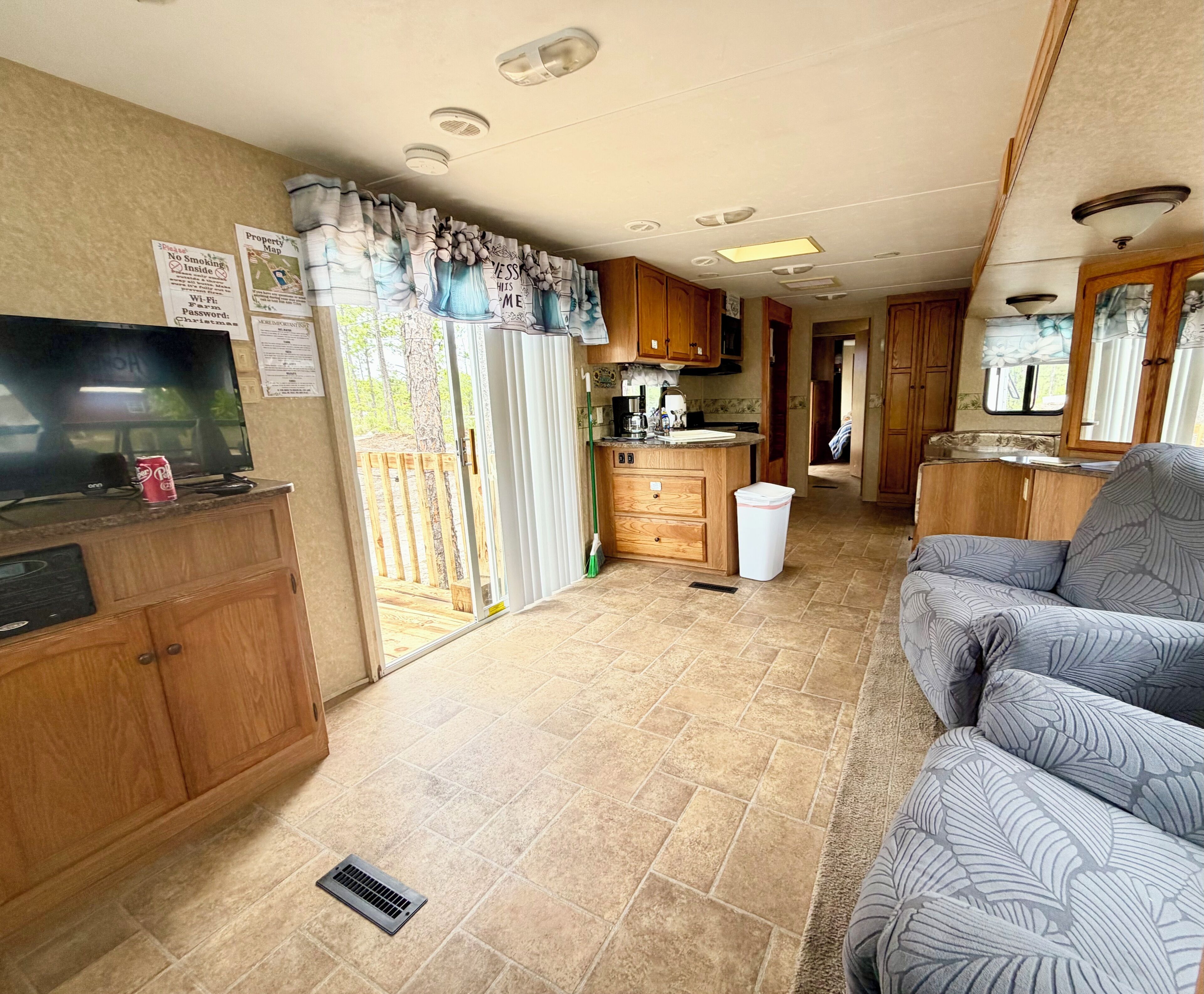 A compact RV interior with a kitchenette, a TV, a sofa, and a sliding door leading to an outdoor deck.