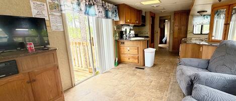 A compact RV interior with a kitchenette, a TV, a sofa, and a sliding door leading to an outdoor deck.