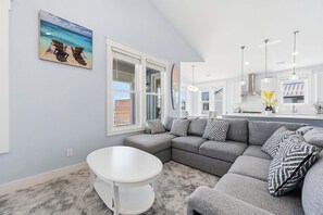 A modern living room with a gray sectional sofa, a round white table, and a painting of chairs on a beach. - Blue Dolphin: Oceanview, game room and Grill (North Topsail Beach)
