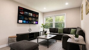 Un salon moderne doté d’un téléviseur à écran plat diffusant divers services de streaming, d’un canapé gris, d’une table basse et d’une grande fenêtre.