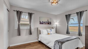 A bedroom with a bed, curtains, a cityscape painting, a floor lamp, and a view of the outdoors. - Modern 4BD Home in Lake Hills Pet friendly (Lake in the Hills)
