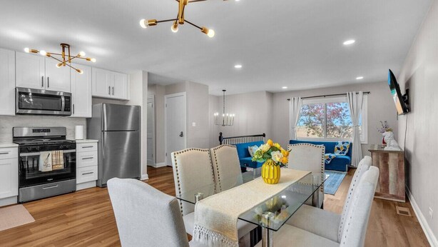 A modern kitchen with stainless steel appliances, a dining area with a glass table and cushioned chairs, and a living area with a blue sofa and a window view. - Modern 4BD Home in Lake Hills Pet friendly (Lake in the Hills)