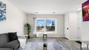 Een moderne woonkamer met een grijze bank, een klein tafeltje met een plant, een groot raam met uitzicht en een deur.