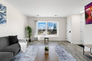 A modern living room with a grey sofa, a small table with a plant, a large window with a view, and a door. - Westchester Corporate Stay 5 Beds (Westchester)