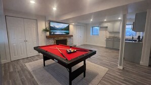 Uma mesa de bilhar com superfície vermelha, uma televisão montada acima dela e uma área de cozinha visível ao fundo.