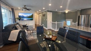 Uma sala de estar moderna com uma mesa de jantar de vidro, assentos confortáveis, uma TV na parede e uma cozinha com eletrodomésticos de aço inoxidável.