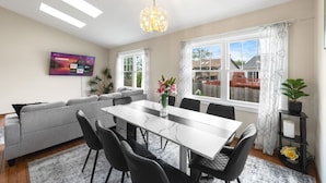 A modern living room with a grey sofa, a dining table with chairs, a TV mounted on the wall, and a large window with curtains. - Family Haven in Streamwood | Ideal for Stay Long (Streamwood)