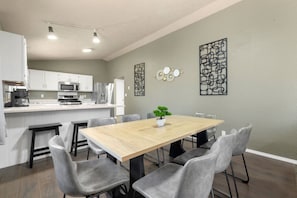A dining area with a wooden table, grey upholstered chairs, a small potted plant, and a kitchen in the background. - 5BR Furnished | Insurance Ready | 7 Beds (Rockford)