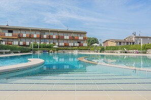 Piscina com deck ladrilhado, cadeiras de descanso e um edifício com varandas ao fundo.
