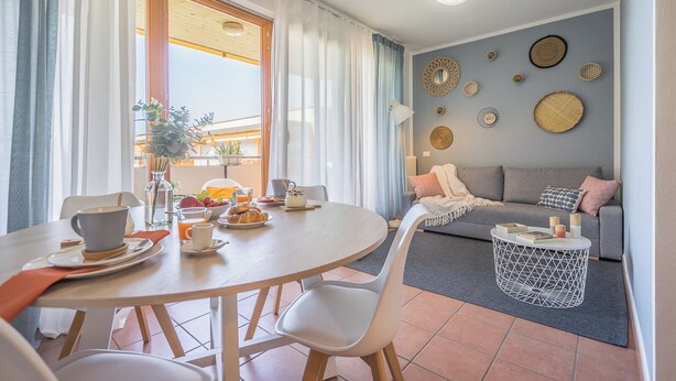 Ein Essbereich mit einem runden Tisch, Stühlen und Frühstücksgebäck. Im Hintergrund befinden sich eine Couch und ein Couchtisch.