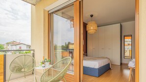 Un balcó amb cadires de vimet, una taula petita amb una ampolla i vasos, i un llit amb matalàs blau a l'habitació propera.