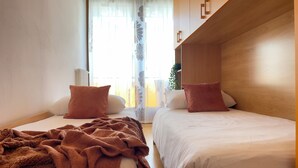A room with two beds, a wooden headboard, a window with sheer curtains, and a radiator. - Luminoso Appartamento con piscina secondo piano (Sirmione)