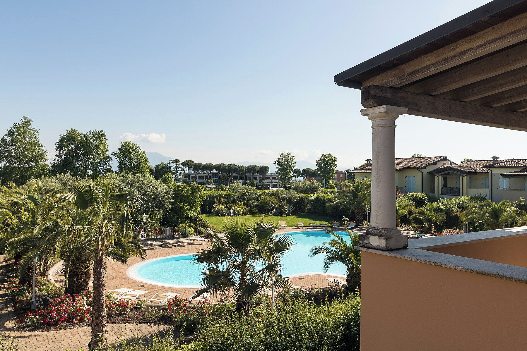 Una piscina con lettini, circondata da vegetazione e con edifici sullo sfondo.