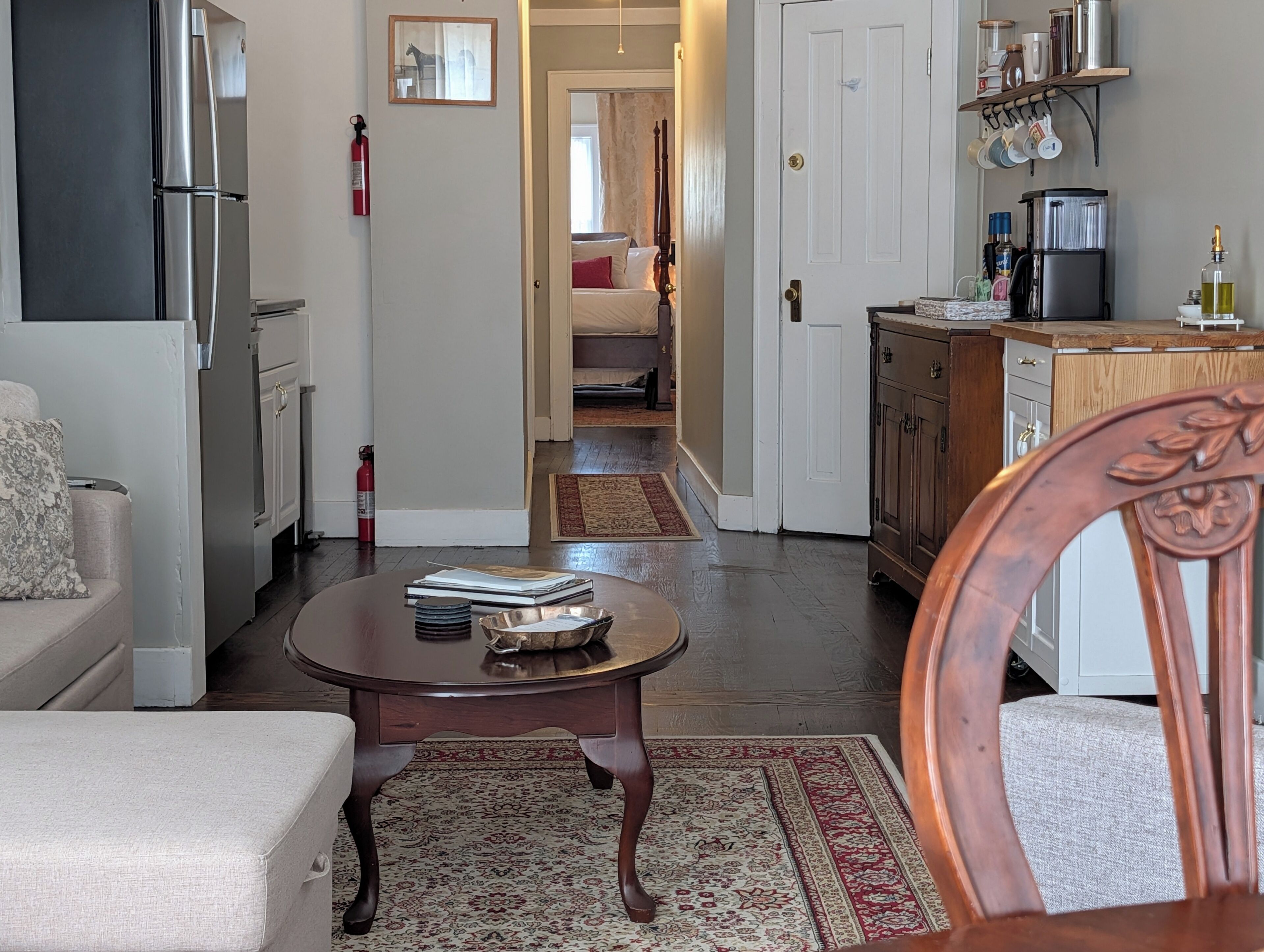 Cocina moderna con refrigerador, estufa y cafetera. Cuenta con una zona de comedor con mesa y sillas, y un pasillo que conduce a un dormitorio.