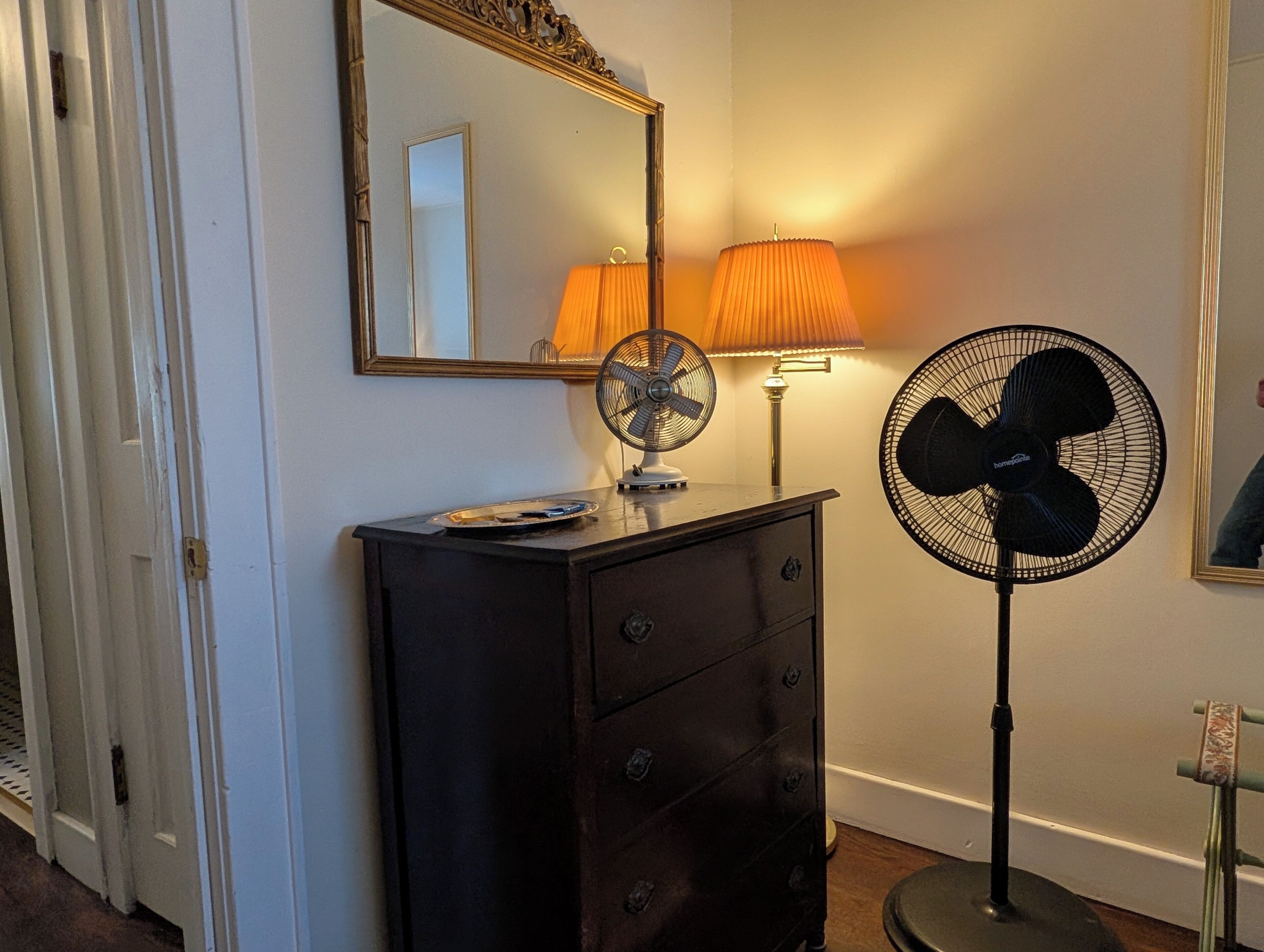 Ein Zimmer mit einem Schminktisch, einem Standventilator, einer Lampe, einem Spiegel und einer Tür.