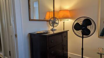 Ein Zimmer mit einem Schminktisch, einem Standventilator, einer Lampe, einem Spiegel und einer Tür.