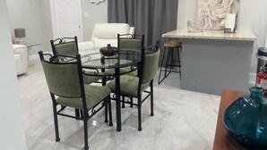 Un coin repas avec une table en verre et quatre chaises, un canapé blanc, un rideau gris, une horloge et un îlot de cuisine agrémenté d’un vase.