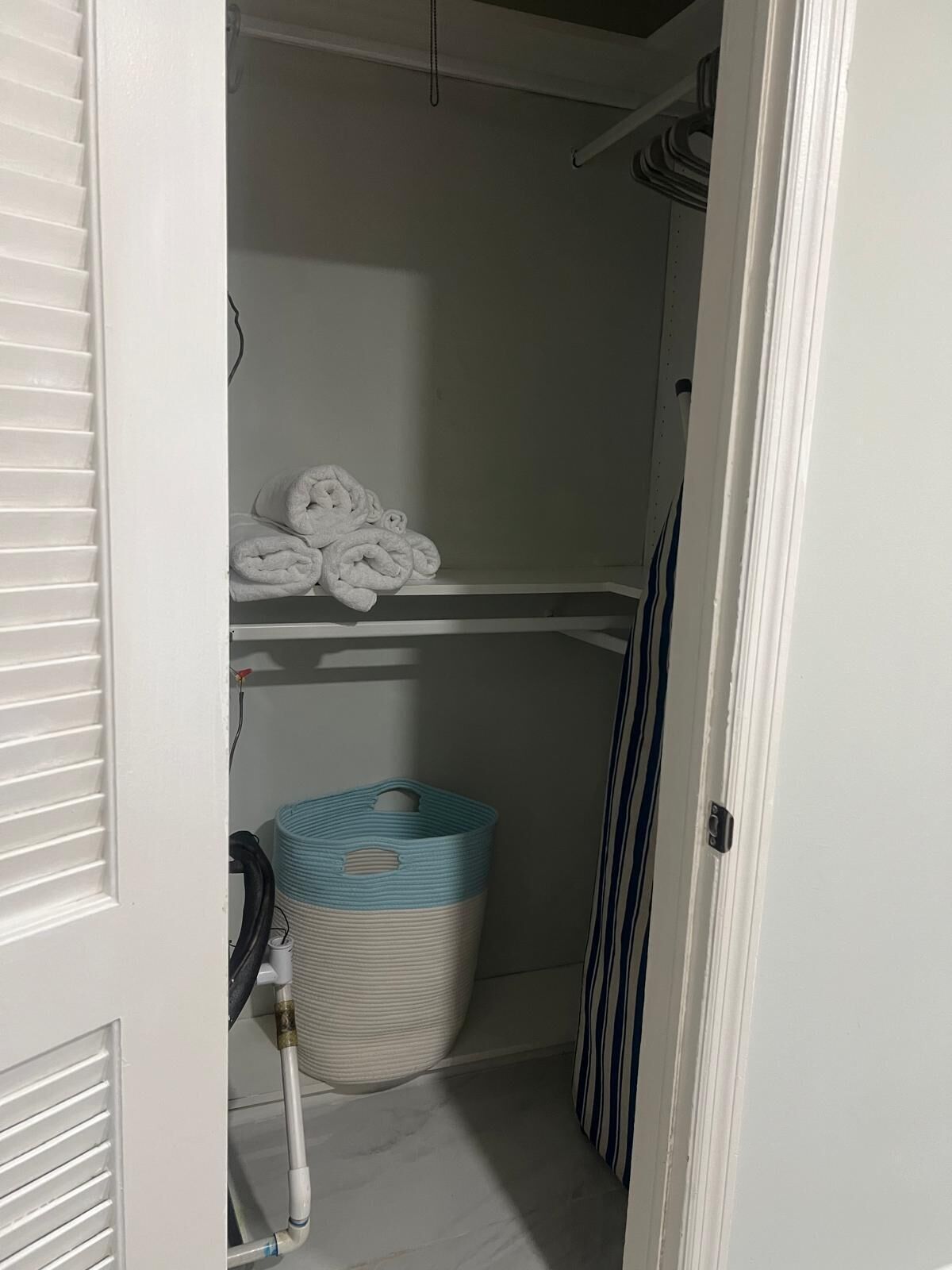 Un armario pequeño y cerrado con una puerta blanca, un perchero, una cesta de ropa a rayas y un tubo blanco.