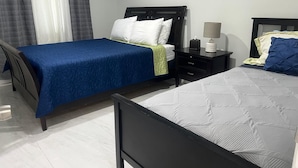 Un lit double avec une couvre-lit bleue et une housse de couette blanche matelassée, une table de chevet avec une lampe et une fenêtre avec des rideaux.