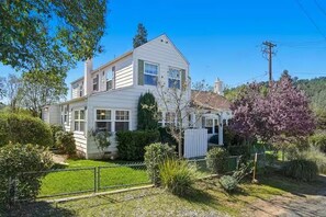 Une maison à deux étages avec un jardin devant, une zone clôturée et un arbre aux fleurs violettes.