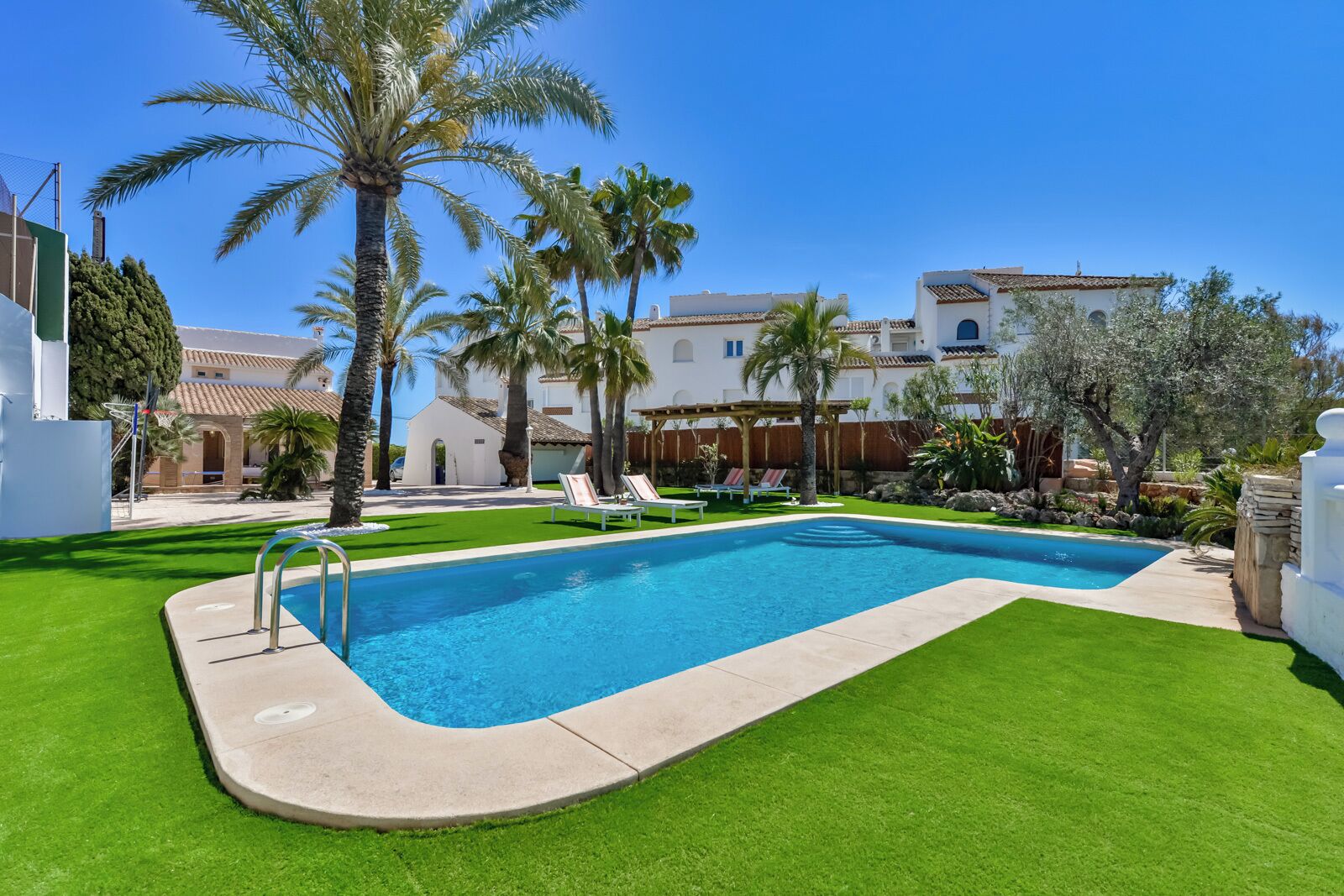 A swimming pool with a surrounding lawn, palm trees, and a building with a tiled roof.