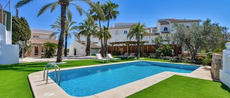 A swimming pool with a surrounding lawn, palm trees, and a building with a tiled roof.