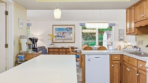 Una cocina con armarios de madera, lavavajillas, zona de comedor con sillas y ventana con vistas a vegetación.