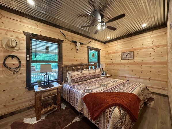 A log cabin-style bedroom with a bed, a nightstand, a lamp, a window with blinds, and a ceiling fan.
