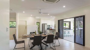 Una cocina moderna con armarios blancos, un comedor con mesa y sillas, y un amplio ventanal corredizo que da a un espacio exterior.