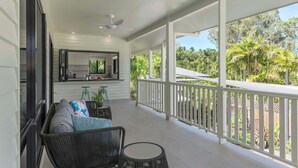 Un porche techado con ventilador de techo, una zona de estar con sofá y sillones, una cocina con mostrador y vistas a exuberante vegetación.