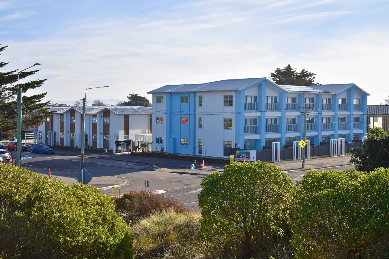 Um edifício de vários andares com fachada azul e branca, uma faixa de pedestres e um estacionamento com carros.