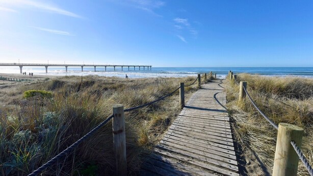 En träpromenad som leder till en strand, med en brygga synlig i bakgrunden.