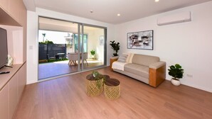 A modern living room with a sofa, a coffee table, and a TV. There is a sliding glass door leading to an outdoor patio. - Coastal 1BR Retreat – Steps to New Brighton Beach (Christchurch)
