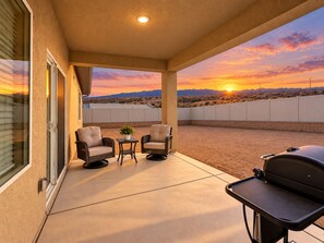 A covered patio with two chairs and a small table, a barbecue grill, and a view of a sunset over a desert landscape.