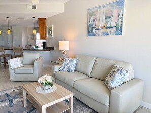 A living room with a sofa, chairs, a coffee table, a lamp, and a painting of sailboats. - Upscale Beachfront Condo • Gulf View Balcony • Pool• Steps to Johns Pass &Dining (Madeira Beach)