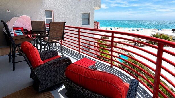 A balcony with wicker furniture, a table, and chairs, overlooking a beach with umbrellas and a pool. - Upscale Beachfront Condo • Gulf View Balcony • Pool• Steps to Johns Pass &Dining (Madeira Beach)